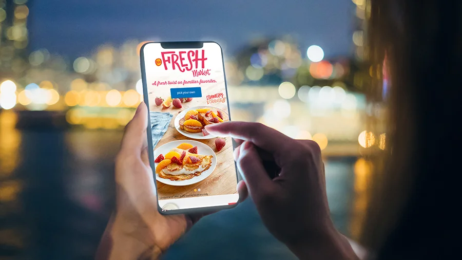 Phone held against city lights showing the Fresh Market mobile page: "A fresh twist on familiar favorites"